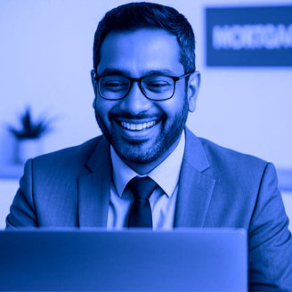 Blue-tinted image of a smiling man wearing glasses and a suit, sitting at a desk and looking at a laptop screen in a professional office setting.