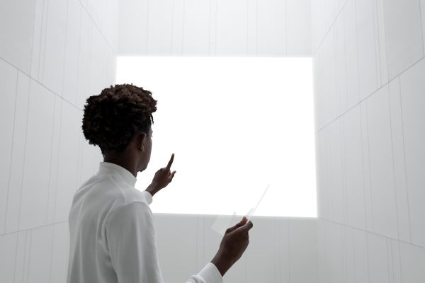 Person standing in a bright, minimal room, gesturing toward a blank white screen or wall, suggesting presentation, planning, or explanation.